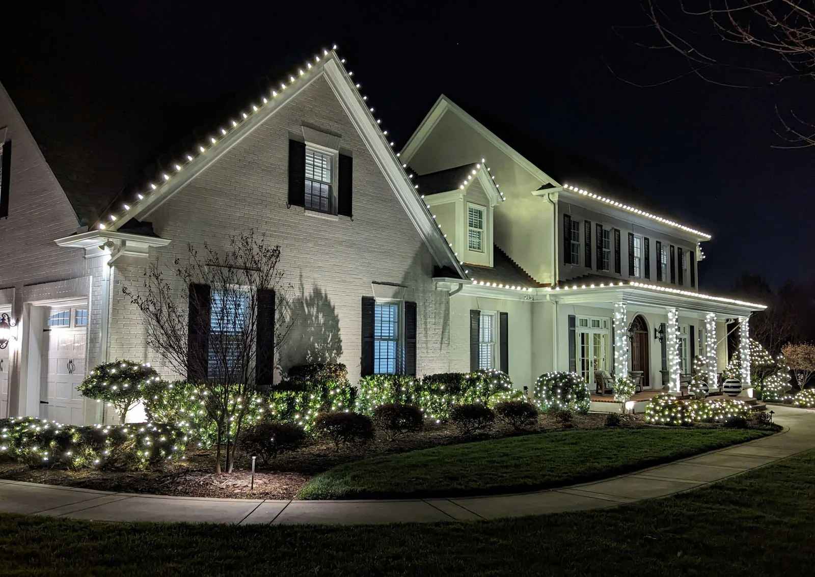 Two-story white brick home at night with white roofline lights and lit shrubs